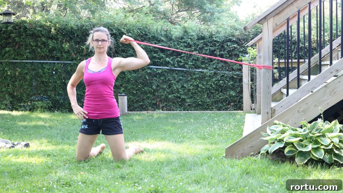 10 Targeted Exercises for Bat Wings + a 10-Minute Arm Routine 7 Woman in pink shirt and black shorts performing a bicep curl exercise with a resistance band outside