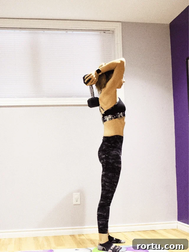 10 Targeted Exercises for Bat Wings + a 10-Minute Arm Routine 8 Woman in dark pants and a sports bra performing a triceps exercise with a dumbbell over her head