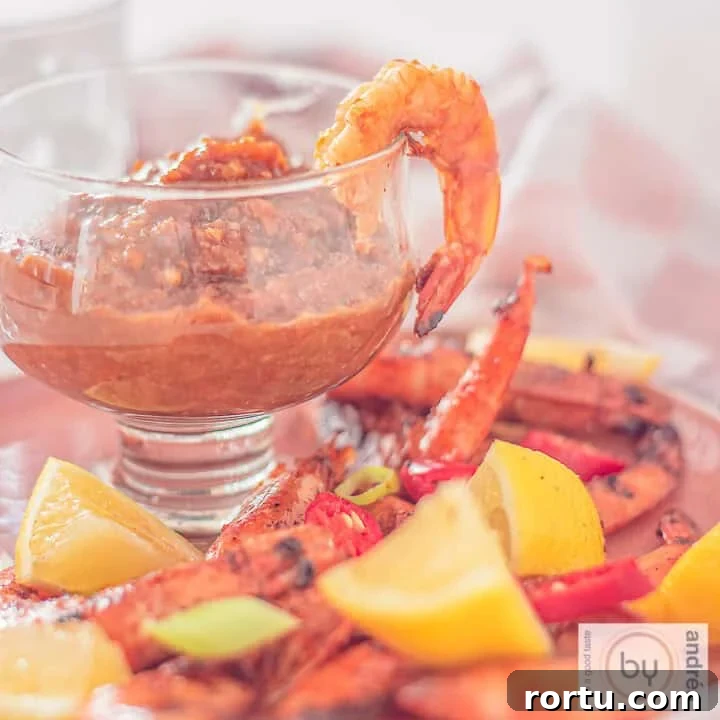 A square photo with a glass filled with cashew satay sauce and grilled shrimps, spring onion, lemon and peppers surrounding it.