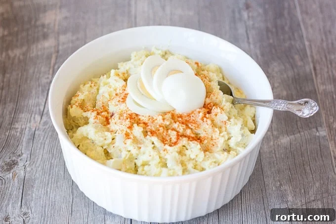 A large white bowl with gluten free potato salad in it and a spoon on the right side. It's topped with a sliced hard boiled egg and some paprika.