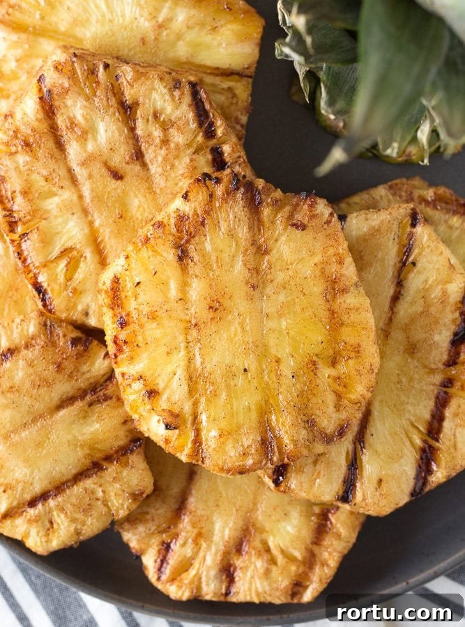 Top down shot of eight grilled pineapple slices on a gray plate with pineapple leaves in the upper right corner.