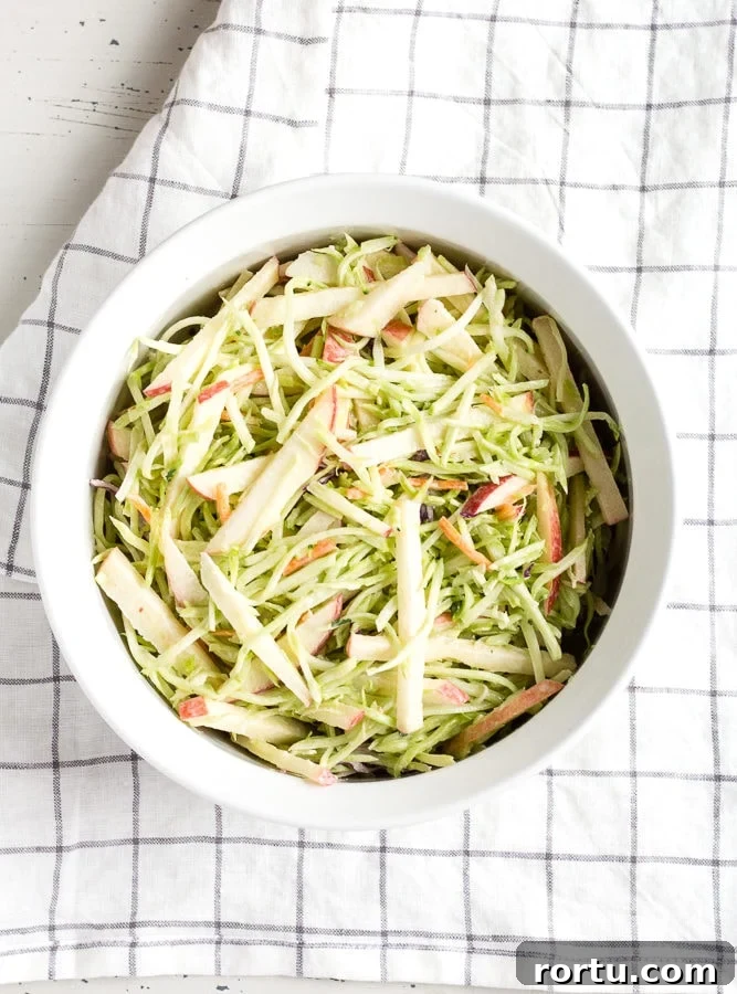 Top down view of apple and broccoli slaw in a white dish with a checked towel underneath.