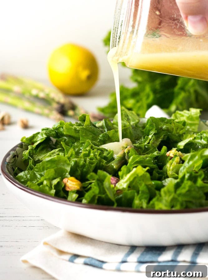 A hand pouring dressing from a mason jar onto spring salad in a low white bowl with asparagus, lemon, and lettuce in the background.