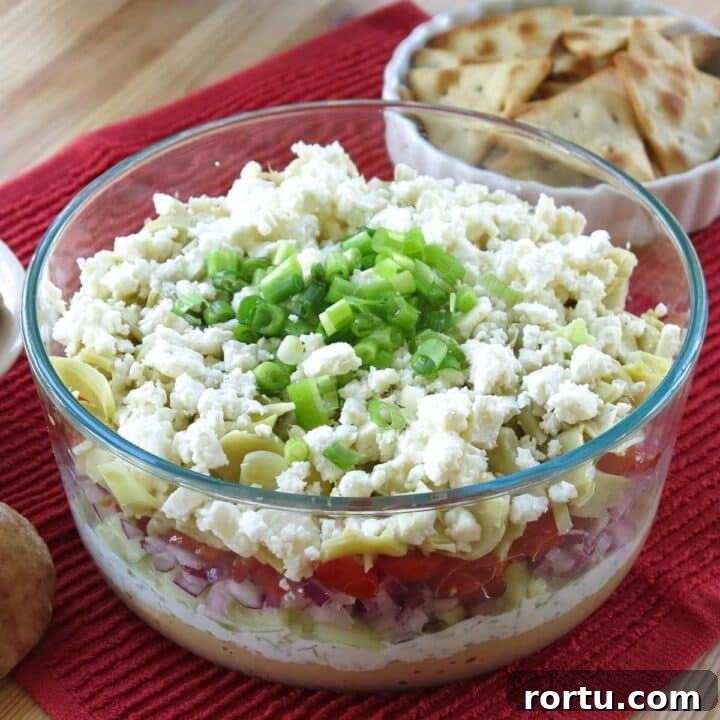 7-layer Greek Dip in a glass bowl with layers of hummus, tsatziki, tomatoes, red onions, feta cheese, and green onions.