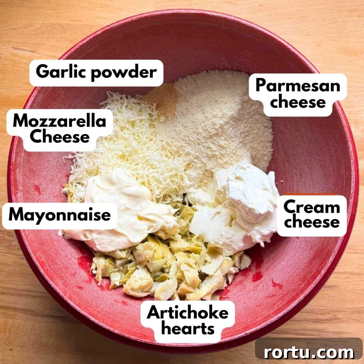 Red bowl with labeled ingredients to make artichoke dip: cream cheese, mozzarella cheese, Parmesan cheese. mayonnaise, chopped artichoke hears, and garlic powder.