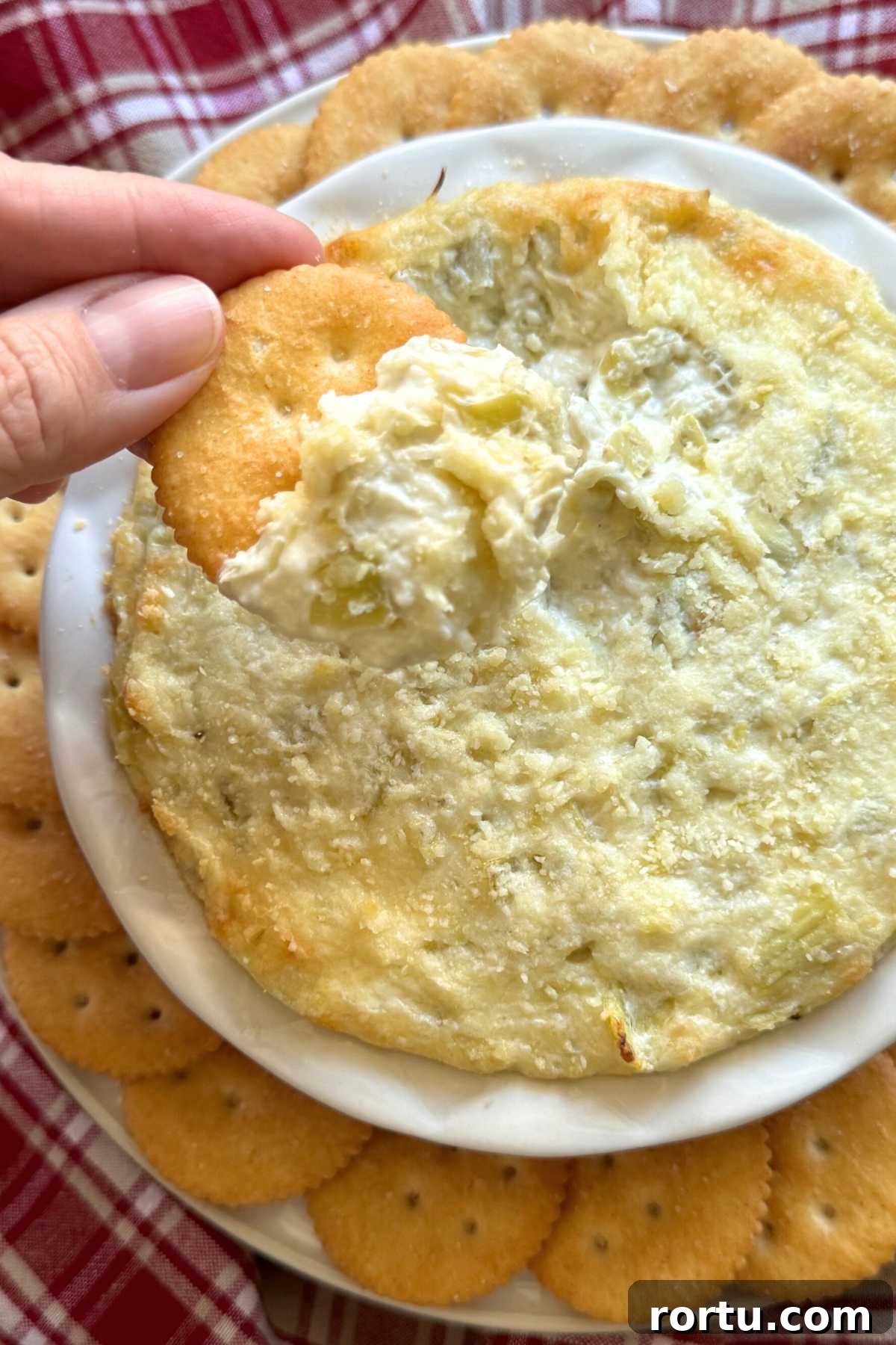 Hand holding a cracker with artichoke dip on it over the bowl of dip with Ritz crackers surrounding it.