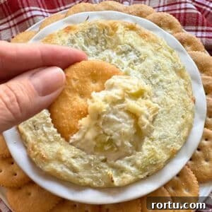 Easy artichoke dip with cream cheese on a cracker over a serving platter with more dip.