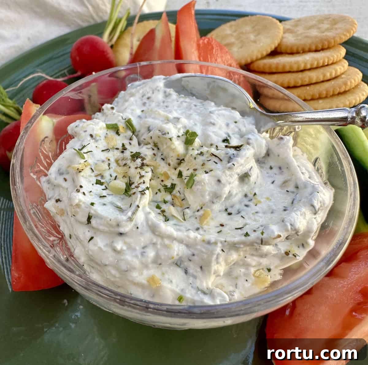 Garlic and herb dip in a dish on a plate with chopped vegetables and crackers.
