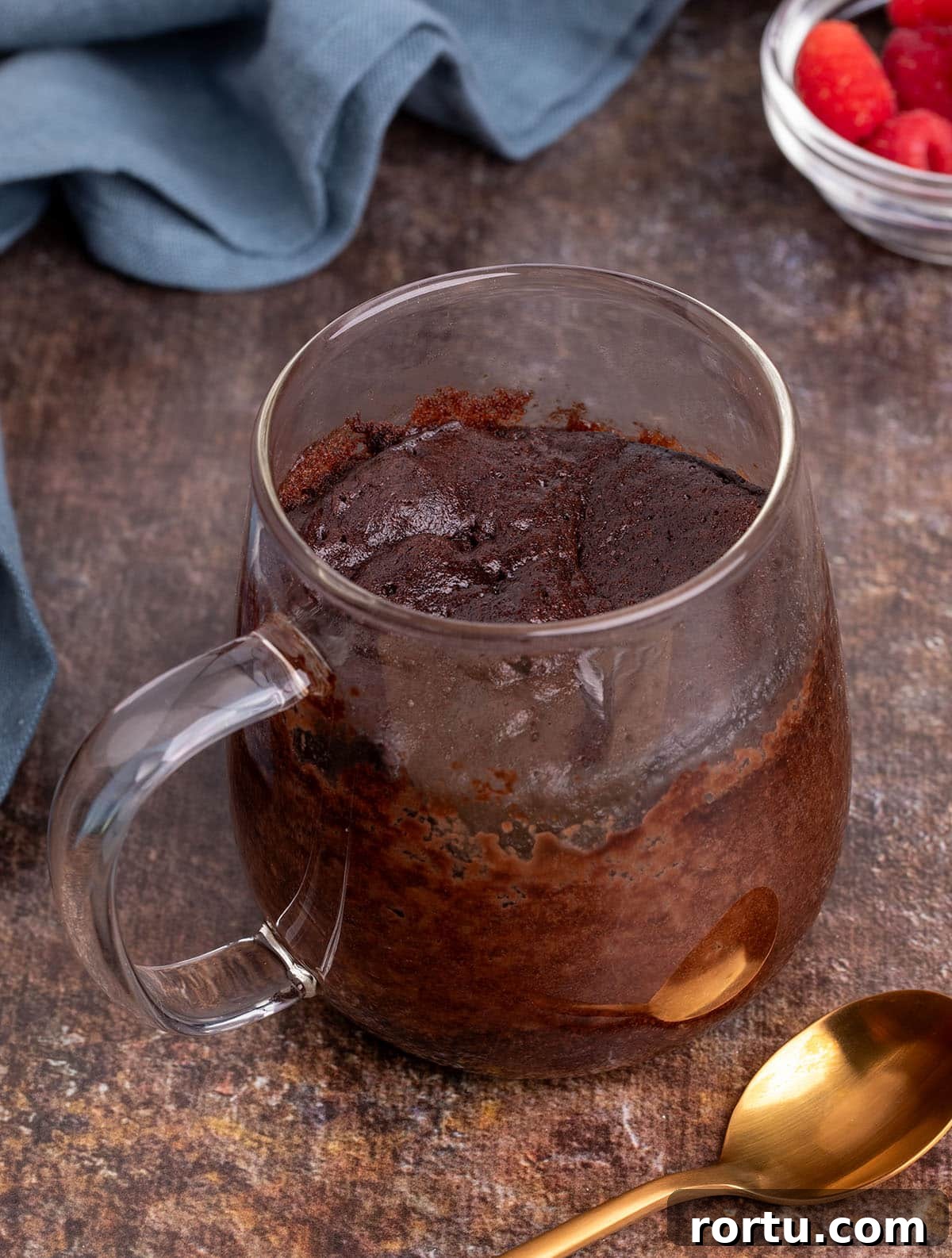 Freshly baked single-serve cake in a clear mug, with a soft, domed top and a gold spoon alongside.