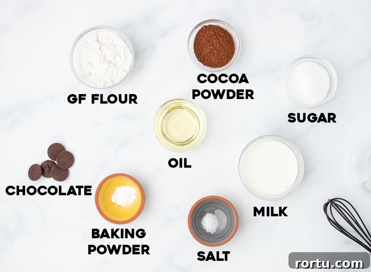 Ingredients for a gluten free mug cake, labeled and arranged on a white surface, including flour, cocoa, chocolate, oil, milk, and more