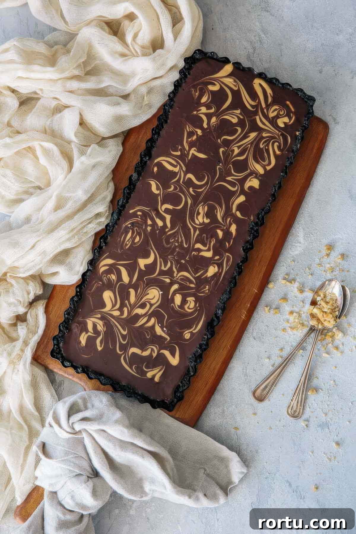 a top-down view of a chocolate ganache tart with a decorative peanut butter swirl on an angle next to a beige towel and two spoons with crushed peanuts