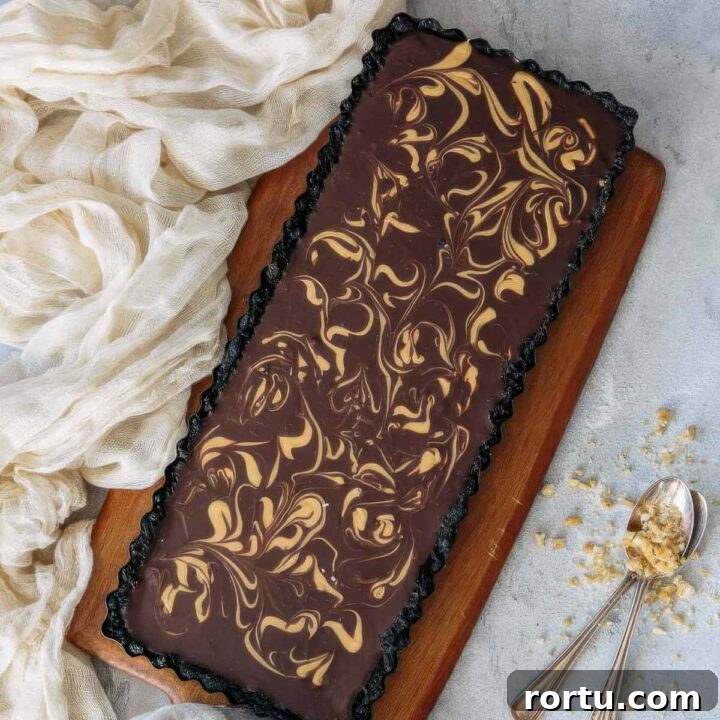 a top-down view of a chocolate ganache tart with a decorative peanut butter swirl on an angle next to a beige towel and two spoons with crushed peanuts