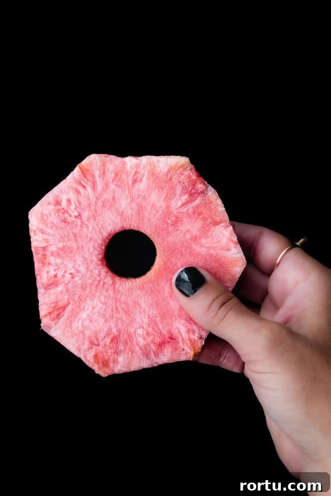 Hand holding a slice of pink pineapple against a black background showing the fruit’s vibrant pink flesh.