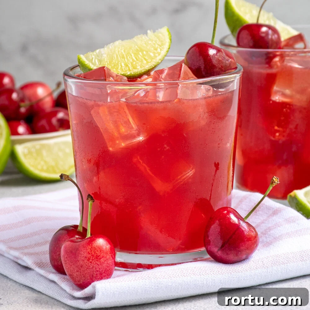 A glass filled with ice and bright red cherry margarita topped with a fresh cherry and lime wedge.
