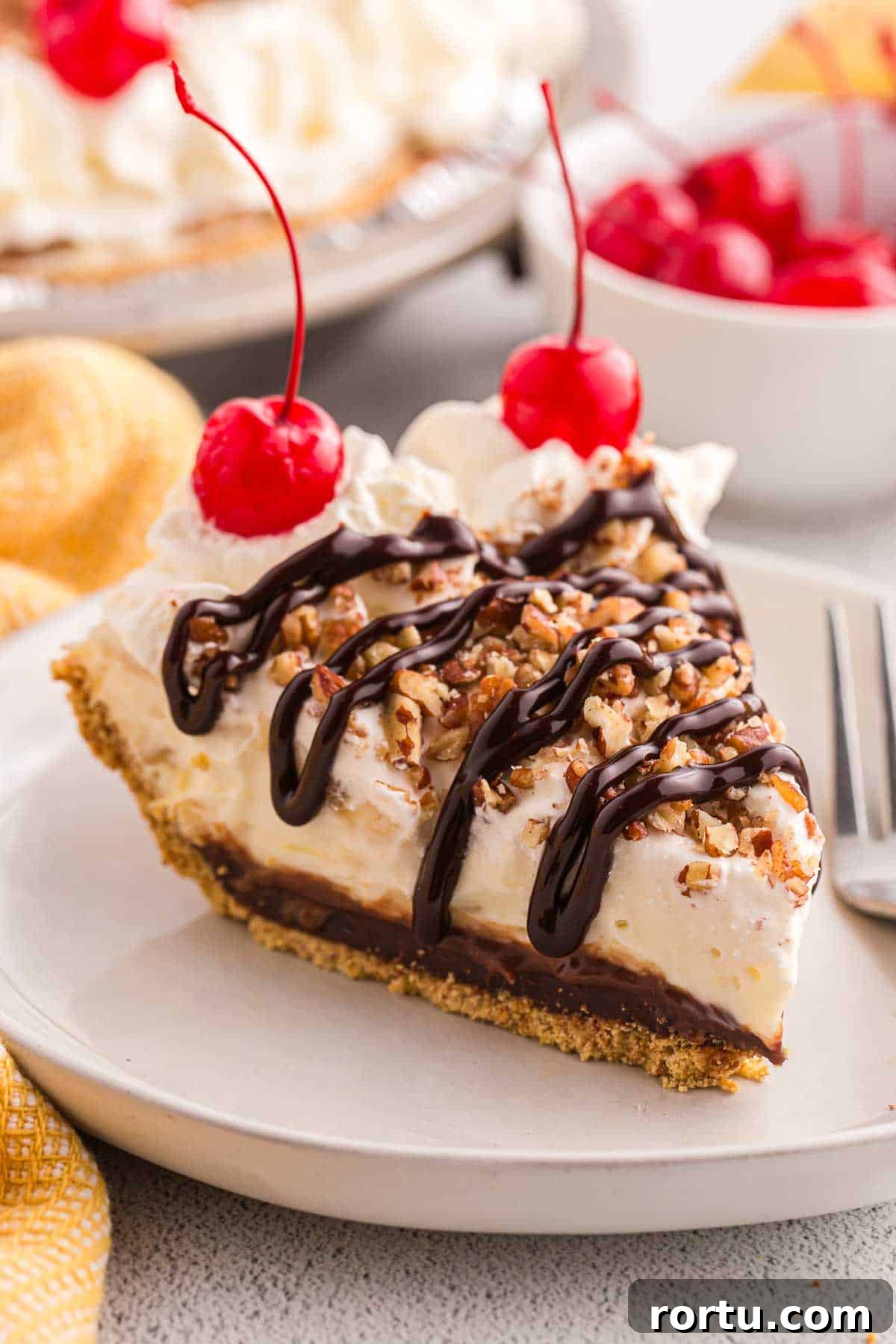Slice of no bake banana split pie topped with whipped cream, chopped pecans, chocolate syrup, and cherries. The slice shows a graham cracker crust with creamy filling and a chocolate layer.