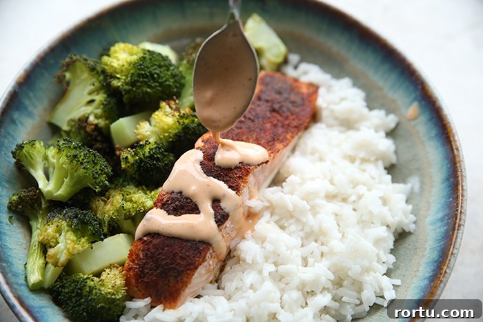A dinner plate with a serving of broccoli, rice and a slice of salmon with a spoon drizzling bang bang sauce on top of it.