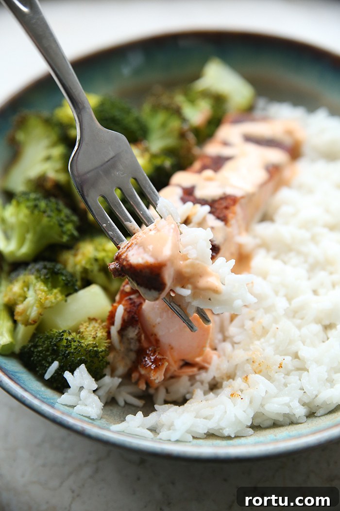 A dinner bowl plate with coconut rice and broccoli and a piece of bang bang salmon in the middle with a fork held above with a piece of salmon on it.