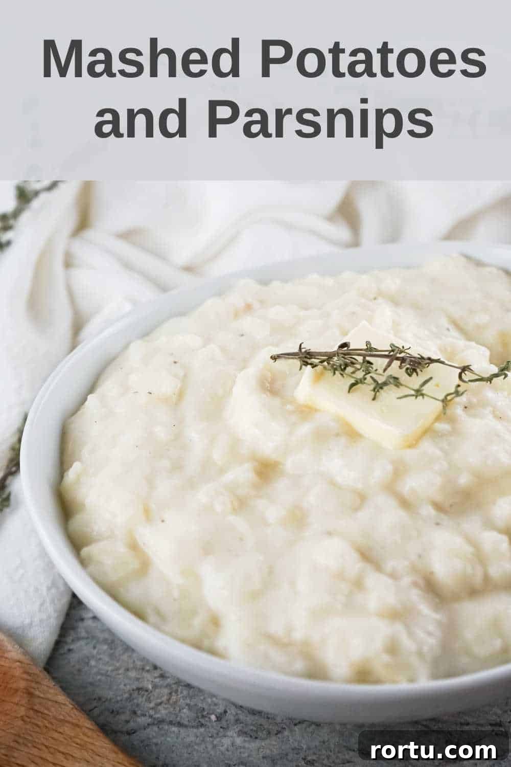 Close up shot of potato and parsnip mash with a pad of butter and herbs in a white bowl on a white cloth.
