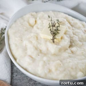 Close up shot of potato and parsnip mash with a pad of butter and herbs in a white bowl on a white cloth.