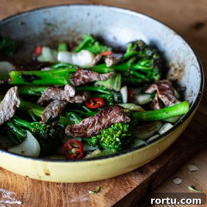 yellow enamel frying pan on wooden surface holding bright green broccolini charred sirloin steak strips and rings of red chilli