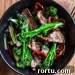 wooden background with black bowl of stir fried broccolini, strips of juicy seared sirloin steak and slices of red chilli