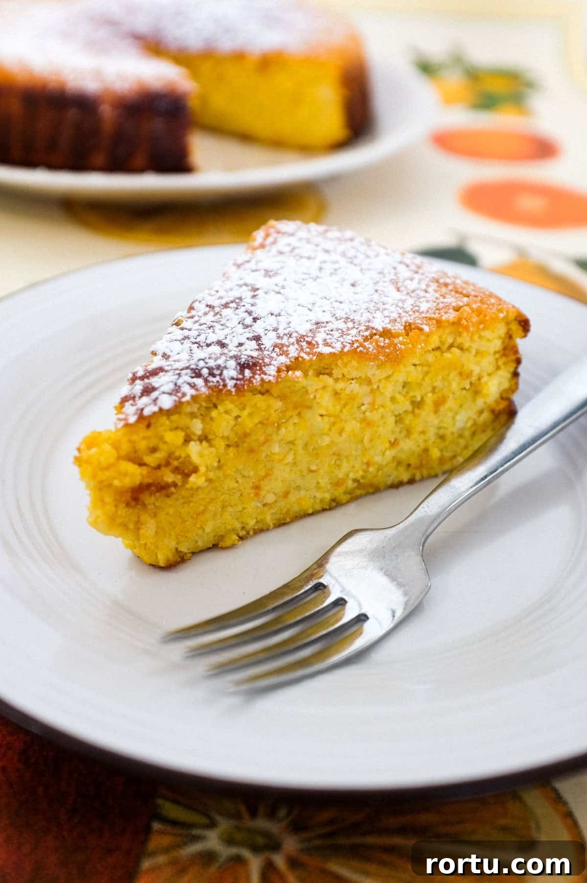 A slice of flourless orange cake sits on a plate with a fork. The rest of the cake is blurred in the background.
