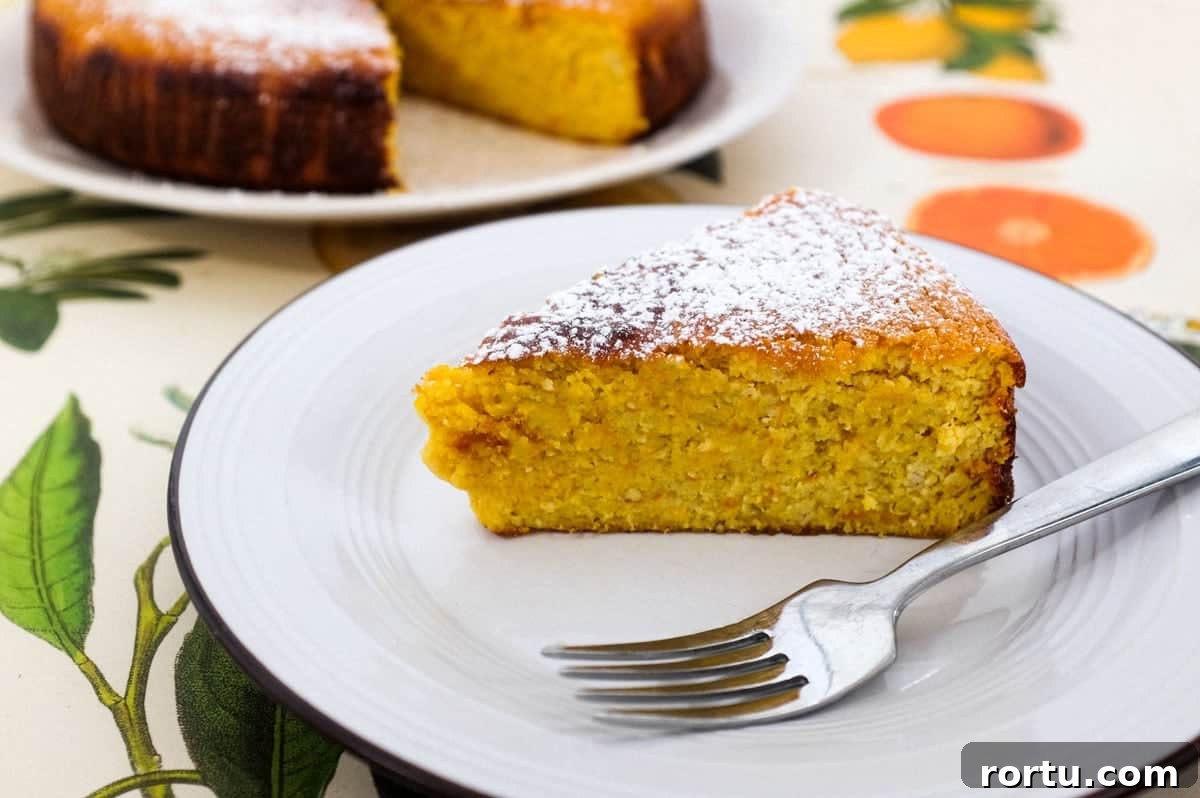 A slice of flourless orange cake sits on a plate with a fork. The rest of the cake is blurred in the background.