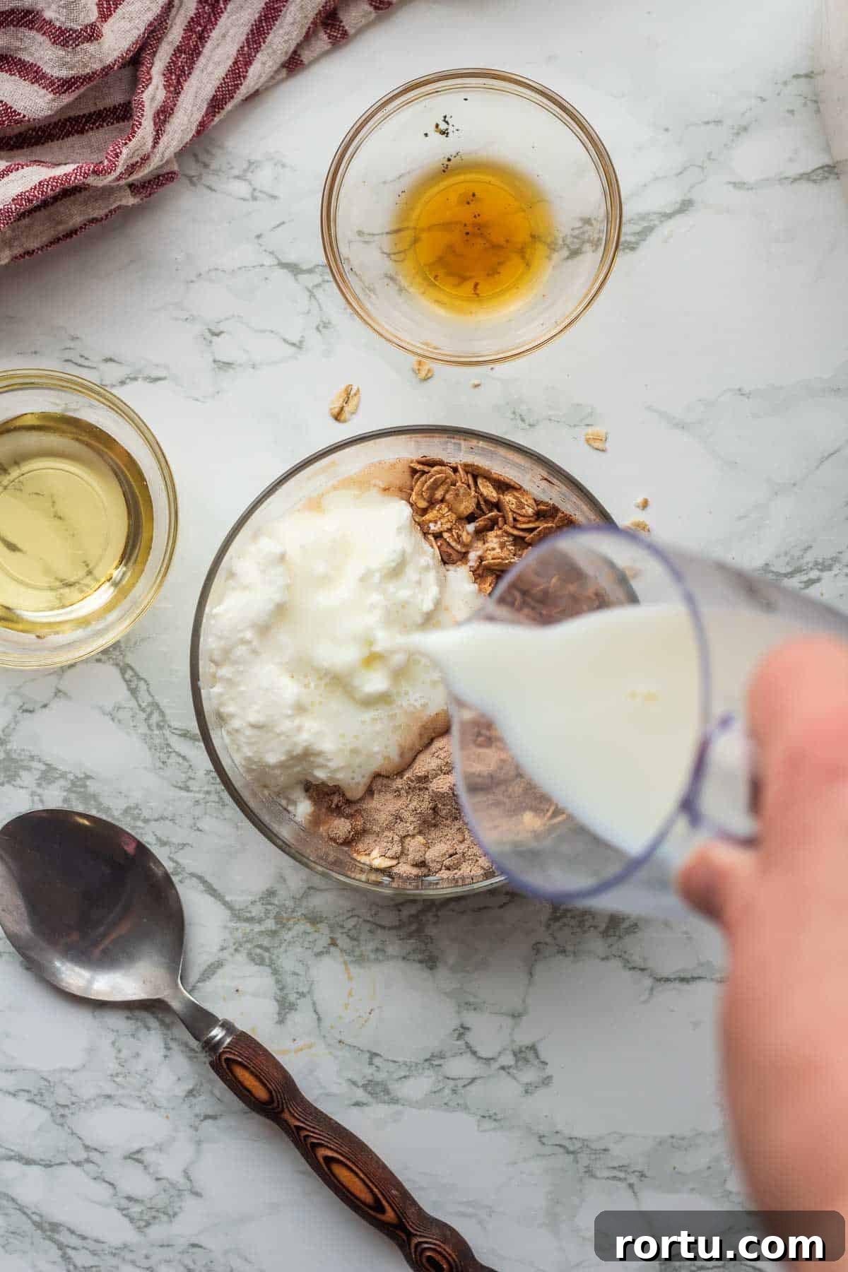 Pouring milk into glass bowl with oats, cocoa powder, protein powder, and greek yogurt.