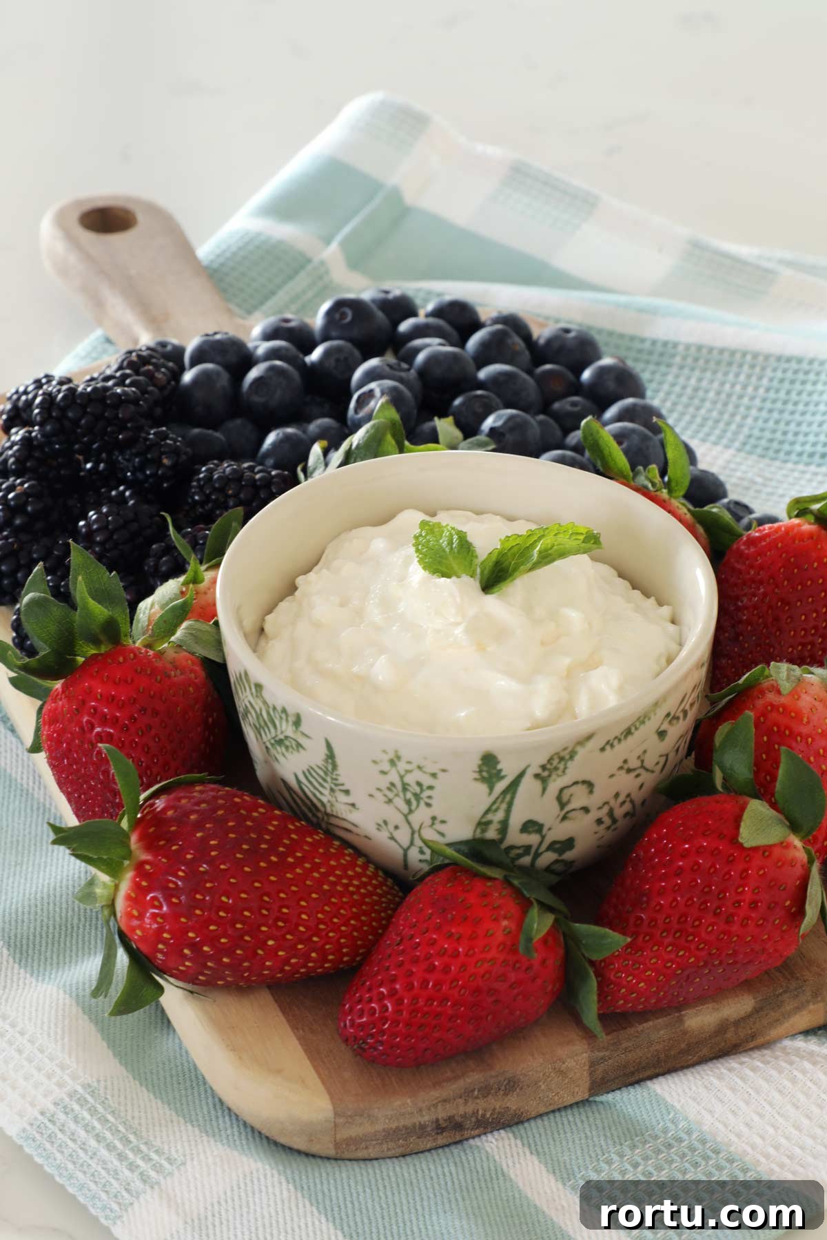 A bowl of fruit dip is on top of a wooden board on top of a teal kitchen towel, strawberries, blueberries and blackberries completely cover the board around the fruit dip bowl.