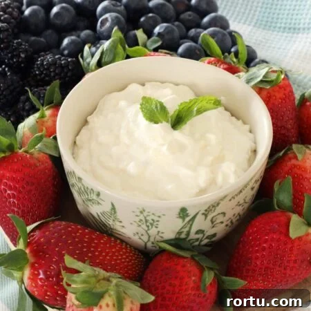 A bowl of fruit dip is on top of a wooden board on top of the kitchen counter, strawberries, blueberries and blackberries completely cover the board around the fruit dip bowl.