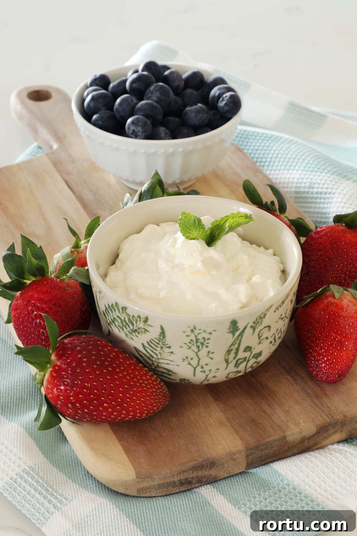 A bowl of fruit dip is on top of a wooden board, strawberries lay on the board while blueberries in a bowl are in the background.