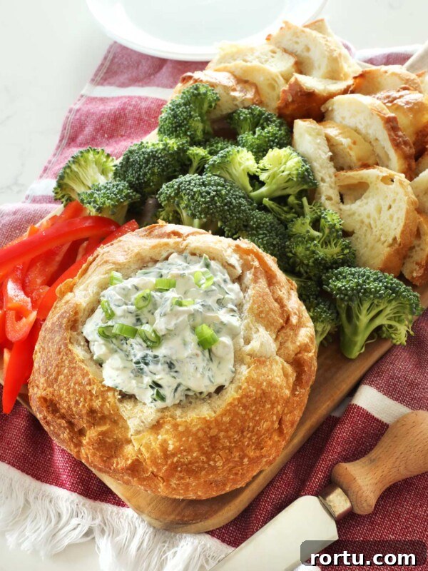 A close up of a bread bowl full of Knorr spinach dip sits on a wooden board along with some broccoli florets, sliced red bell pepper and more bread pieces