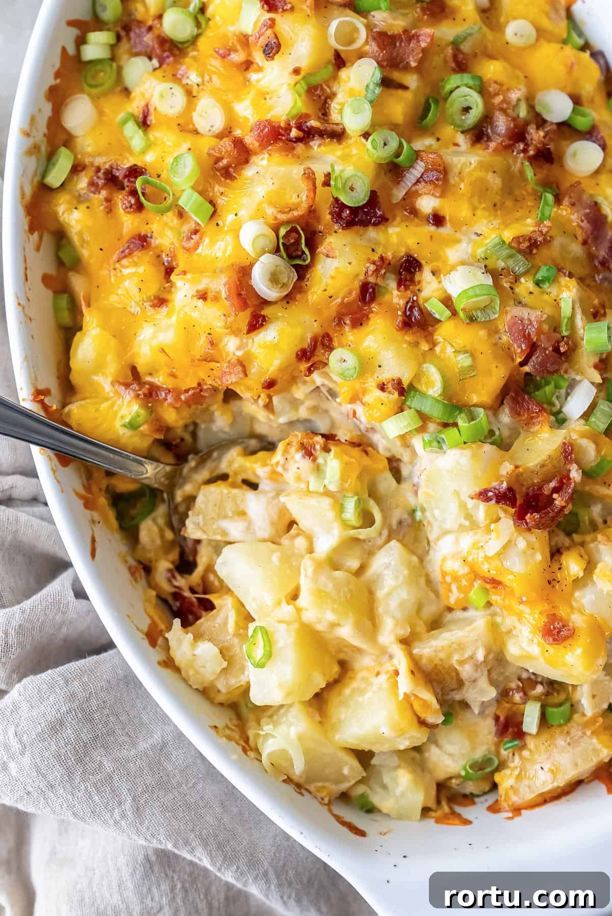 Twice Baked Potato Casserole in a white baking dish with melted cheddar cheese, crispy bacon, and sliced green onions on top. A spoon is scooping out a creamy serving of cheesy potatoes.