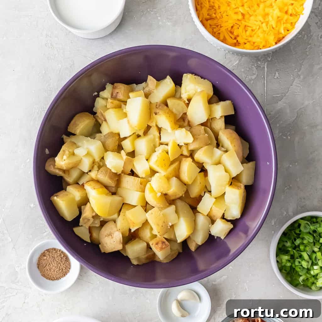 Bowl of chopped baked Yukon Gold potatoes in a large purple mixing bowl with milk, cheese, seasoning, and other ingredients arranged around it.