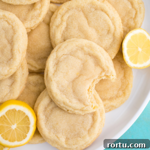 A plate of soft, sugar-coated lemon cookies, with one lemon cookie showing a bite taken out. Slices of fresh lemon are placed beside the delicious lemon sugar cookies on the plate.