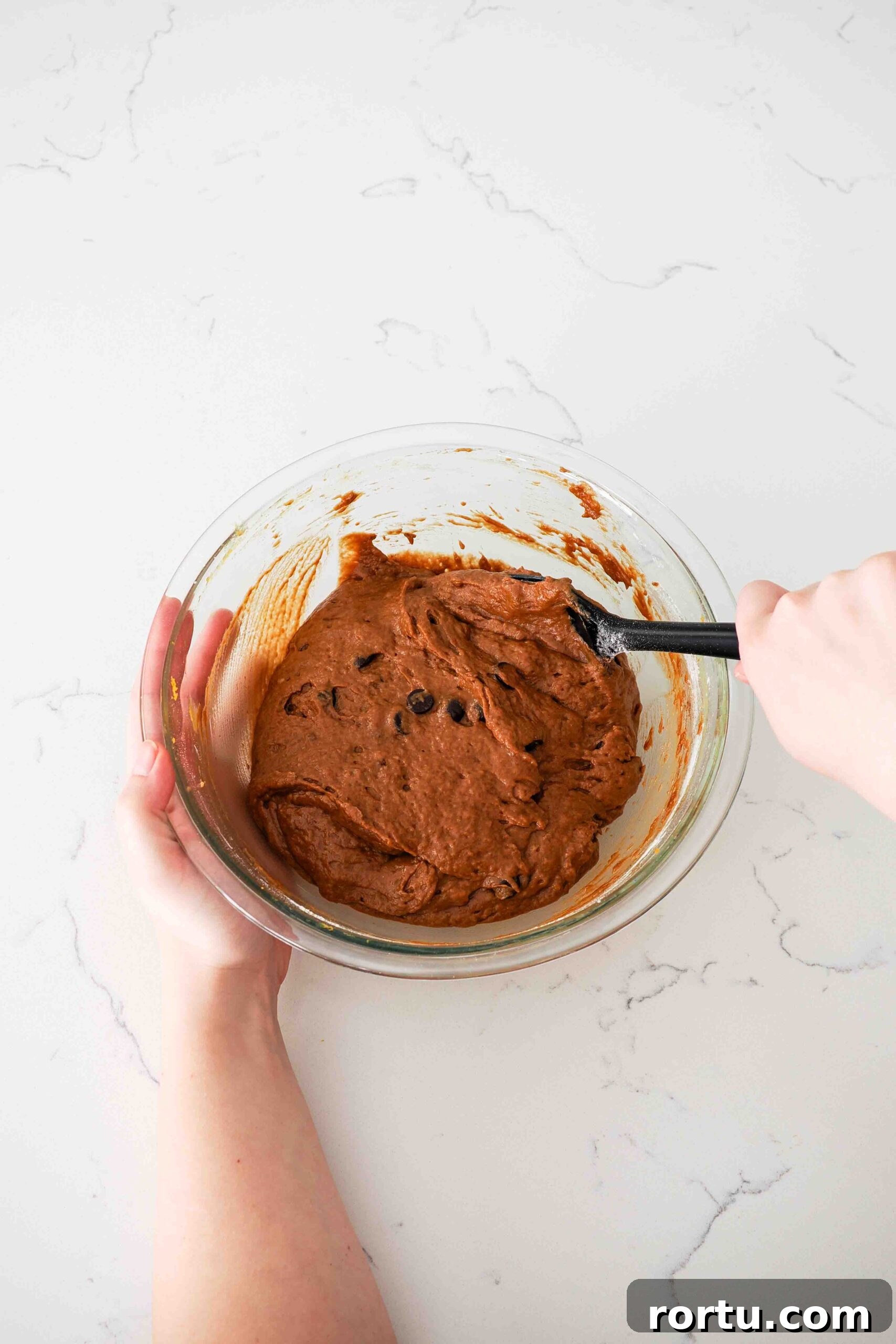 Chocolate Pumpkin Muffins Recipe — Moist Spiced Chocolate Muffins 7 Two hands use a spatula to stir together chocolate pumpkin muffin batter.