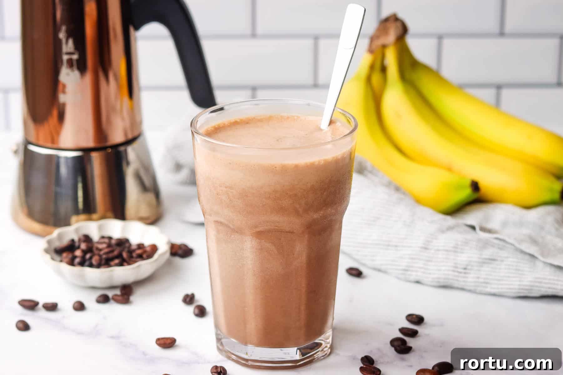 Energizing Banana Coffee Smoothie Recipe for Morning Boost 3 A tall glass of coffee banana smoothie with a spoon in it and coffee and banana in the background.