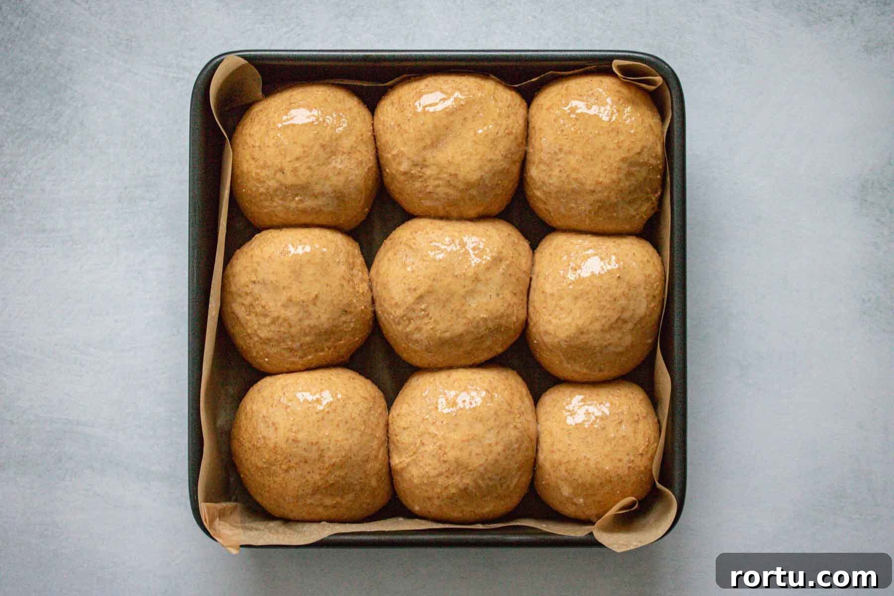 Quick Whole-Wheat Dinner Rolls That Rise Perfectly 10 Rolls on a square baking sheet, topped with molasses and butter, after the second rise, looking puffy.