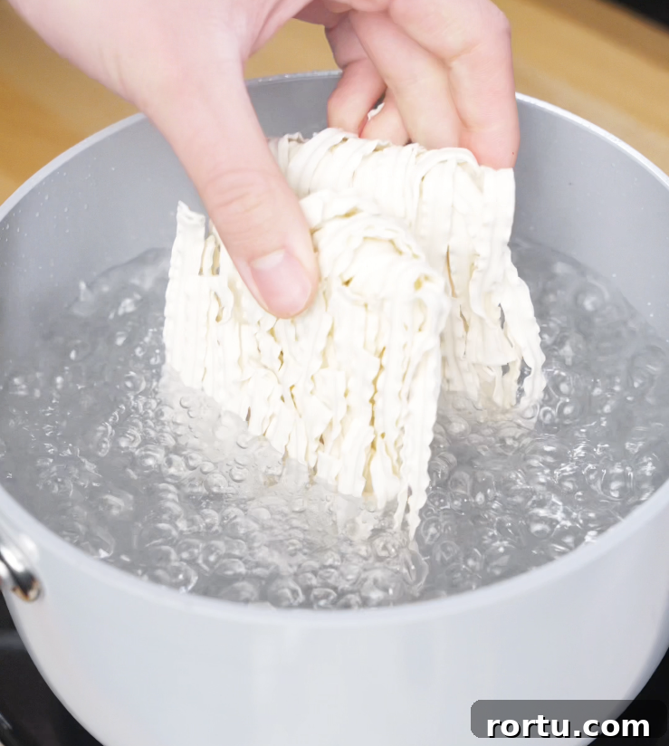 A hand dropping a block of instant noodles into a pot of rapidly boiling water, marking the start of the noodle prep for chili garlic butter noodles.