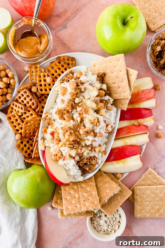 Apple Cinnamon Bites Recipe: Quick No-Bake Snack for Kids and Adults 10 white oval serving dish filled with cream cheese caramel apple dip topped with granola, apple slices and two graham cracker squares surrounded by pretzels, graham crackers, and apples for dipping.