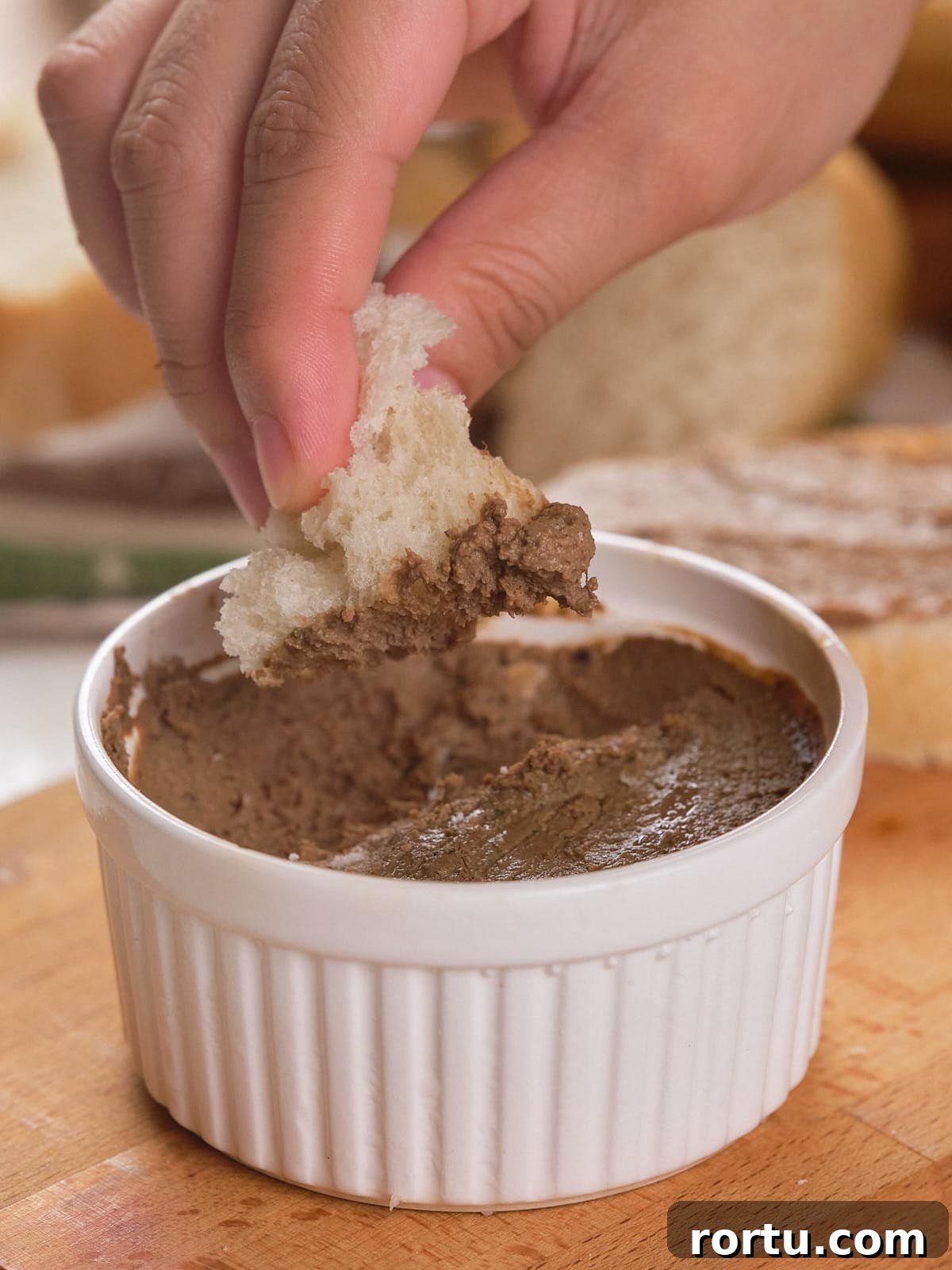 A ramekin of beef liver pate on a wooden board with a hand dipping a bread into it.