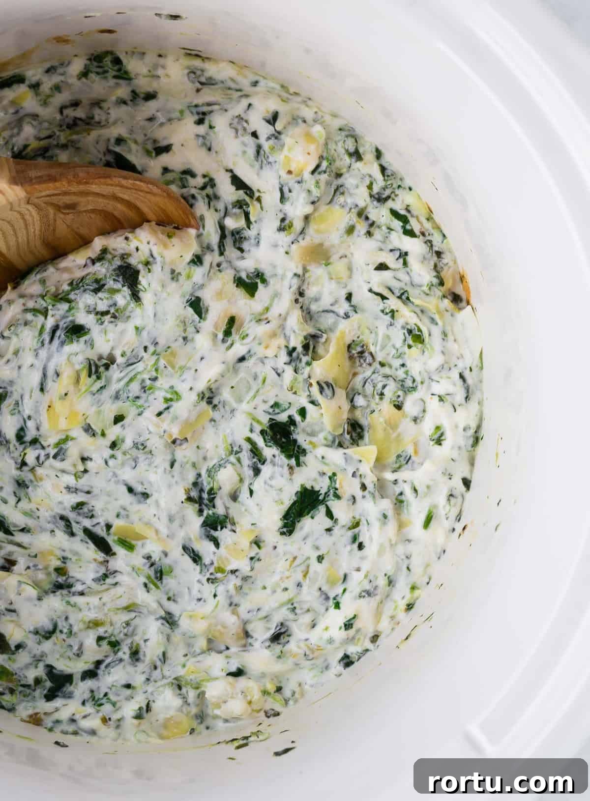 Overhead view of spinach dip with artichoke hearts in a white slow cooker with a wooden spoon.