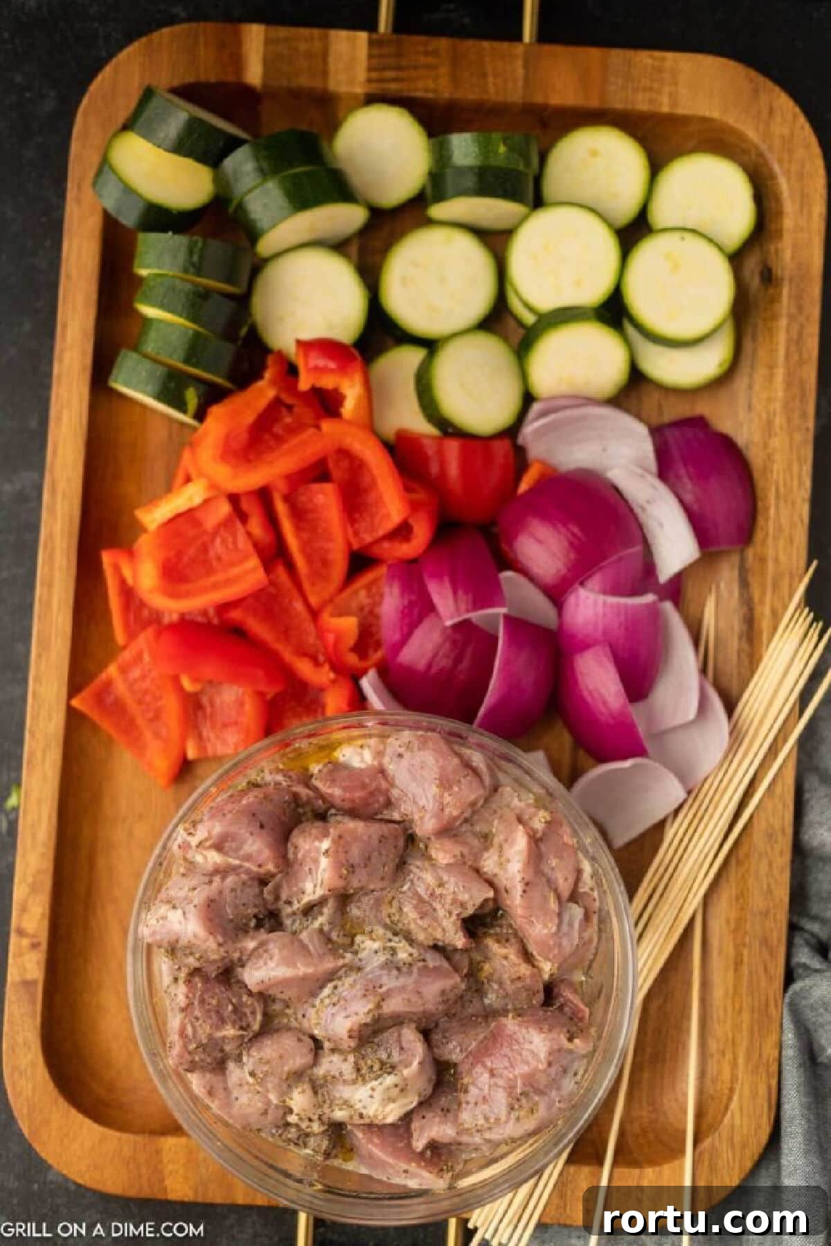 Sliced zucchini, bell peppers, and red onions on a cutting board with a bowl of marinated pork pieces with wooden skewers on the side.