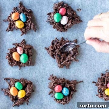 Chocolate noodle nests with candy eggs in the middle on a baking sheet with one nest being shaped with a spoon viewed from above.