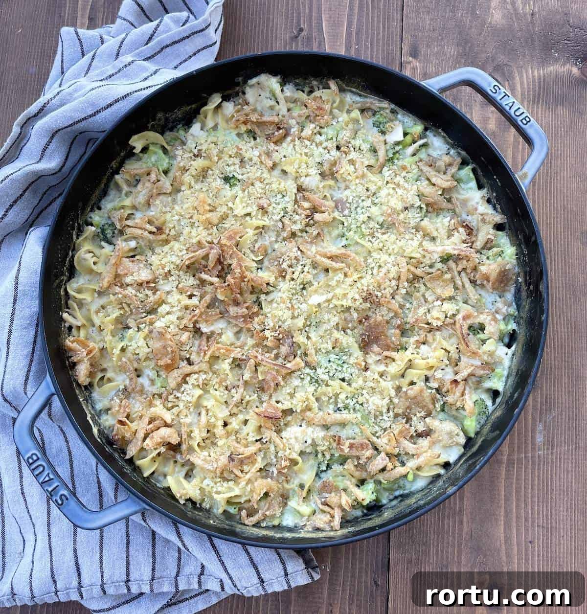 A large cast iron pan filled with cheesy chicken and broccoli noodle casserole on a wooden board.