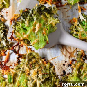 A close up shot of smashed broccoli parmesan on a sheet pan with a spatula lifting up a broccoli piece.