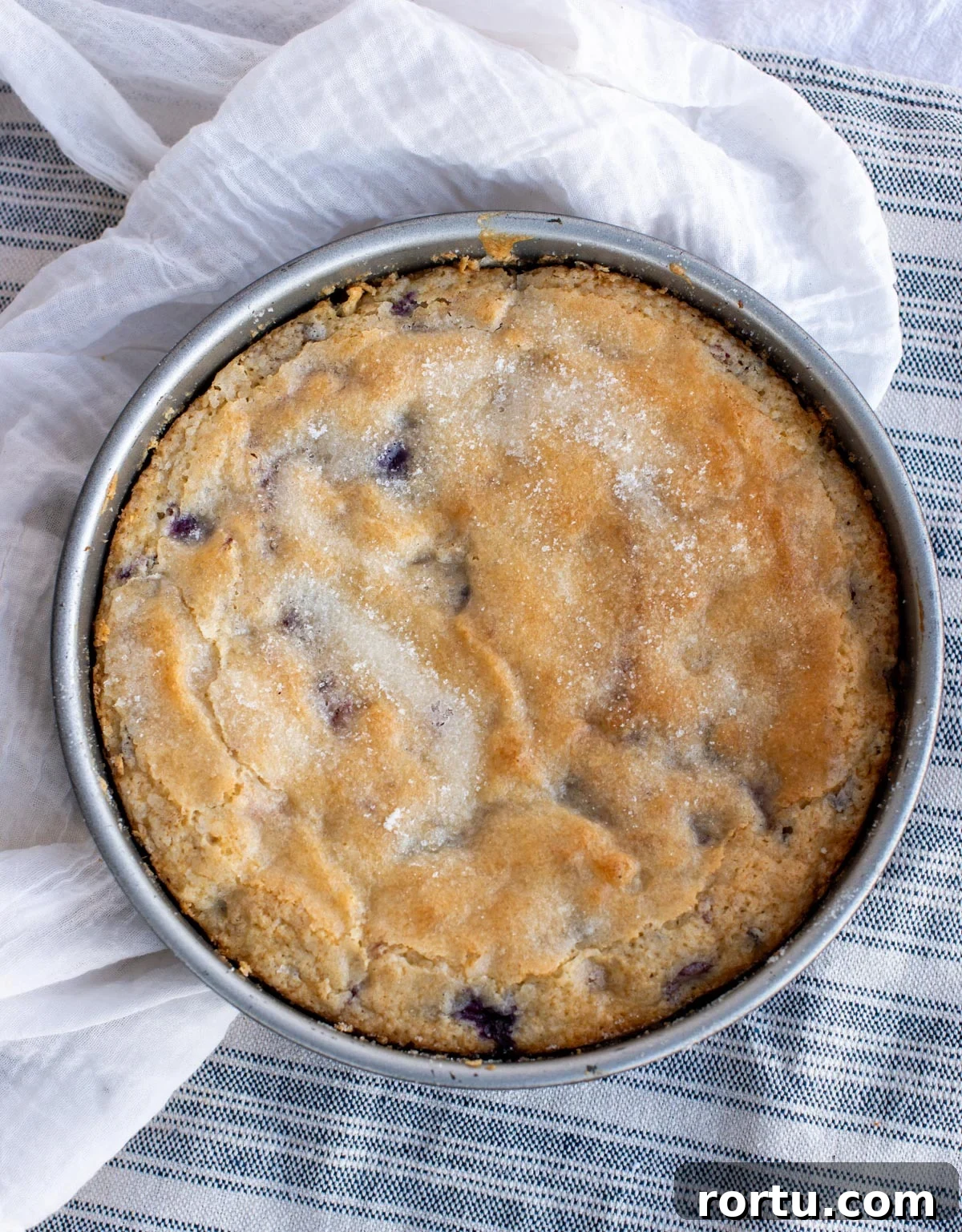 Baked cake with sugar visible on top sits in the cake pan placed on a tea towel.