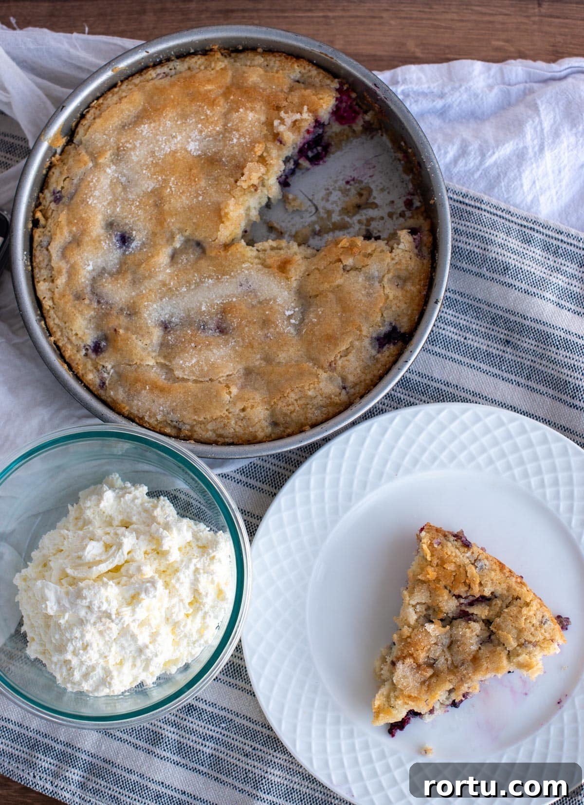 Overhead view of a sourdough discard triple berry cake in a round pan with a slice removed, showing a tender crumb and juicy berries, served with a plate and a bowl of whipped cream.