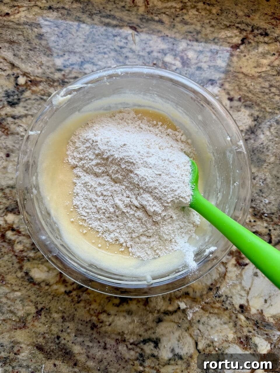 Flour added on top of the wet batter in a glass bowl.