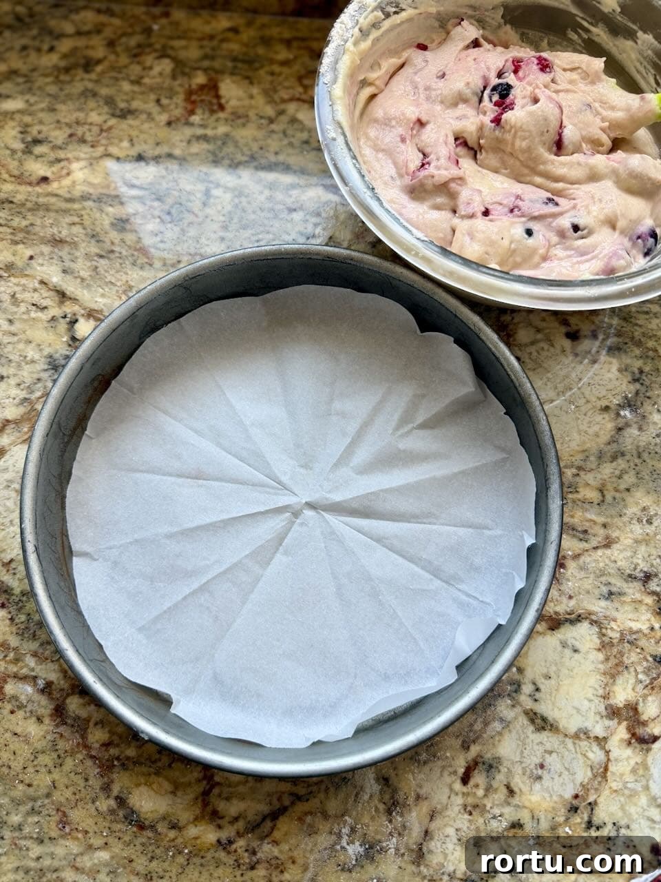 Round cake pan lined with parchment paper next to a bowl of berry cake batter.